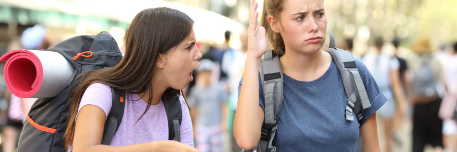 Two young women with backpacks argue outdoors, possibly stressed from travel and not being able to stay fit while on the go.