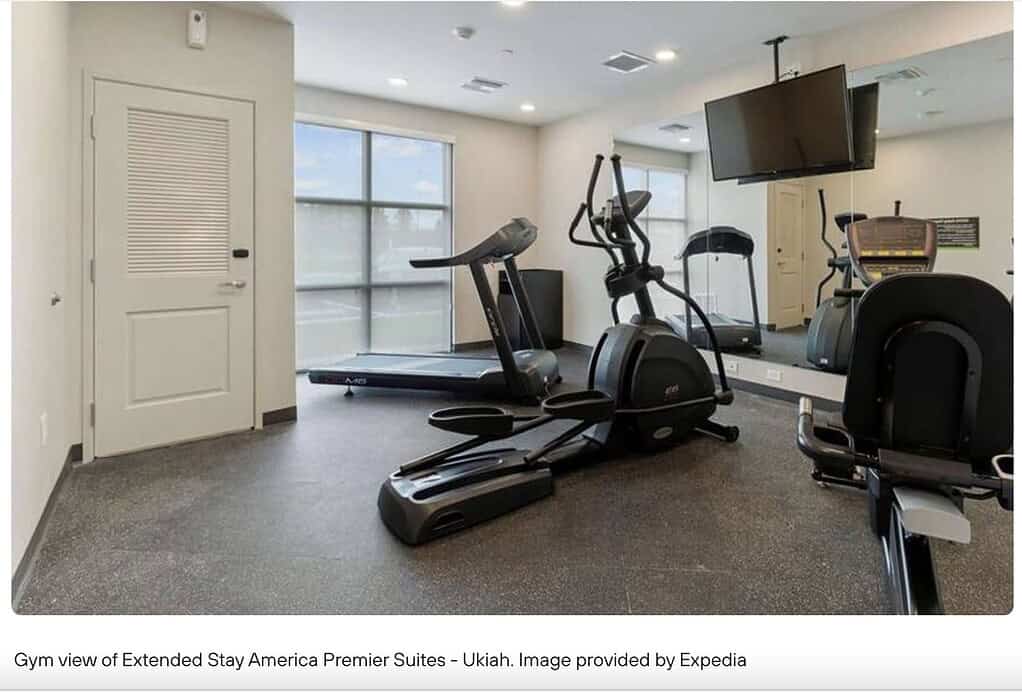A bad example of a hotel gym featuring a treadmill, elliptical, exercise bike, TV, mirrors, and large windows.