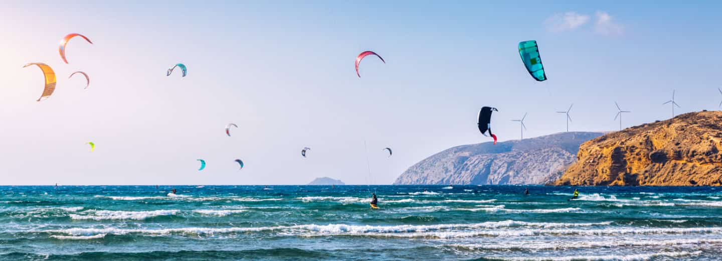 Active kitesurfers ride the waves near a rocky coastline with wind turbines in view, showing fitness and adventure while traveling.