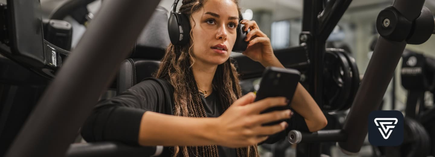 Woman wearing headphones holds a phone while sitting on gym equipment using the Gymverse fitness app.