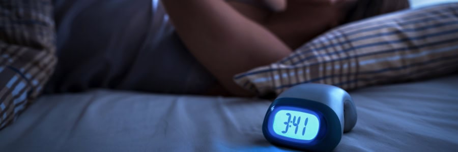 A traveler lies awake in a hotel bed at 3:41 am, digital clock glowing by the bedside, highlighting travel sleep struggles.