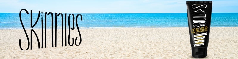 Skinnies sunscreen tube on a sandy beach with ocean and sky, ready for active travelers staying fit during their trips.