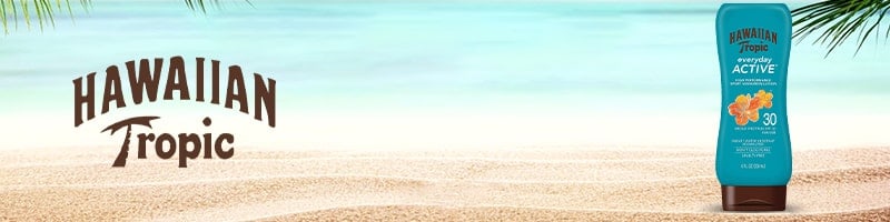 Hawaiian Tropic sunscreen bottle on sandy beach near ocean and palm leaves, perfect for travelers aiming to stay fit outdoors.
