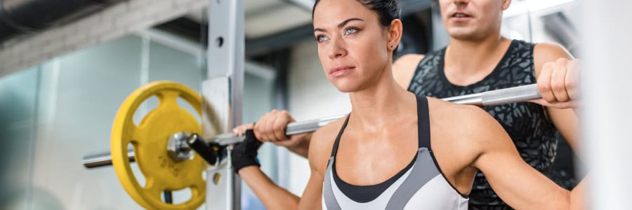 A woman lifts a barbell with yellow weights as a man spots her in a hotel gym, showing how to stay fit while traveling.