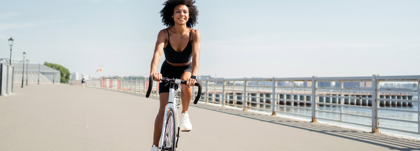 Woman staying fit while traveling, riding a bicycle near a waterfront with city views and railings on a sunny day.