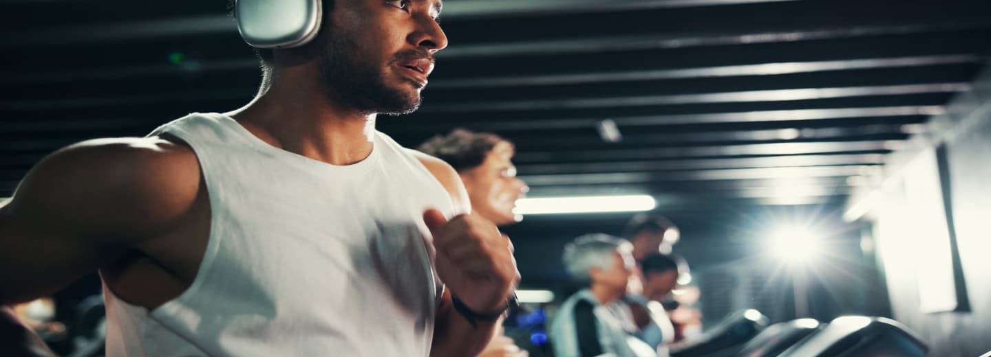 Man in a white tank top runs on a treadmill with headphones in a hotel gym, staying fit while traveling. Others work out nearby.